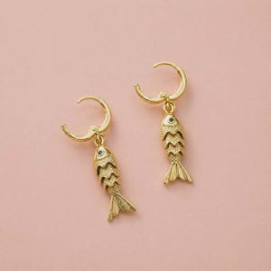 May include: A pair of gold-toned hoop earrings featuring dangling fish-shaped charms. Each fish charm has a textured, layered scale pattern and a small black dot representing an eye. The earrings are designed with a hinged closure mechanism.