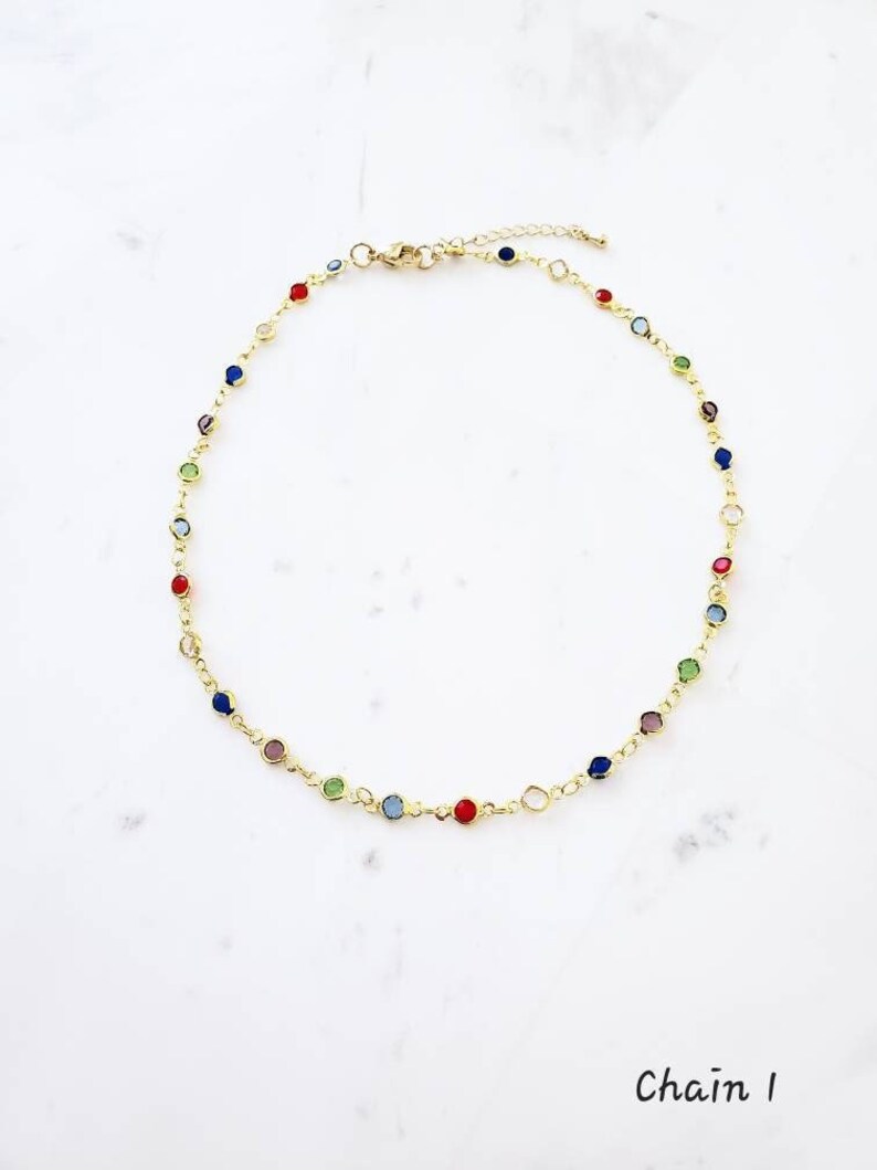 May include: A gold-toned chain necklace featuring a series of small, round, multi-colored gemstones spaced evenly along the links. The necklace includes a lobster claw clasp and an adjustable extender chain. The text "Chain 1" is visible.