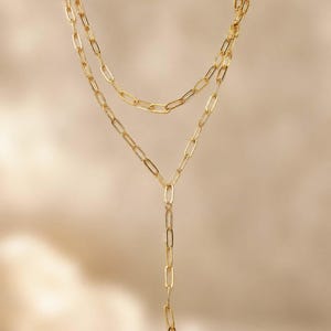 May include: A gold-toned necklace featuring a double-layered design with elongated oval links. The upper layer consists of a shorter chain, while the lower layer forms a Y-shaped drop, with both sections connected by a central vertical chain.