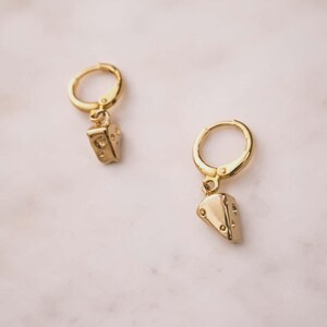 May include: A pair of gold-toned hoop earrings featuring small, dangling charms shaped like triangular wedges of cheese. Each charm has a smooth, polished finish with small, circular indentations scattered across the surface to mimic holes.