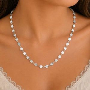 May include: A silver-toned necklace featuring a delicate chain composed of numerous small, flat, circular metallic discs linked together in a continuous, uniform pattern that rests around the neck area.