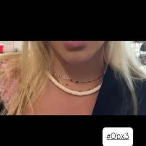 May include: A layered necklace set consisting of two distinct pieces. The top layer is a gold-toned chain featuring small, square-shaped colorful gemstones. The bottom layer is a thick, smooth, off-white beaded strand with a matte finish.