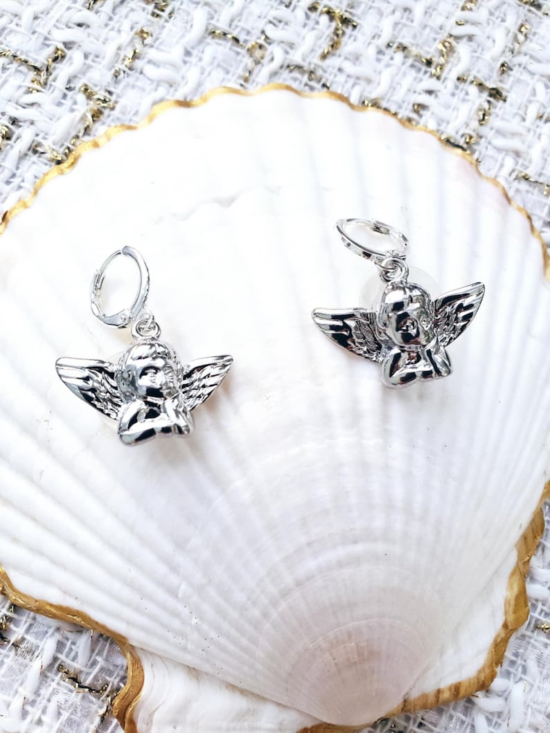 May include: A pair of silver hoop earrings with a small angel charm hanging from each hoop. The angels are in a praying position with their wings spread out.