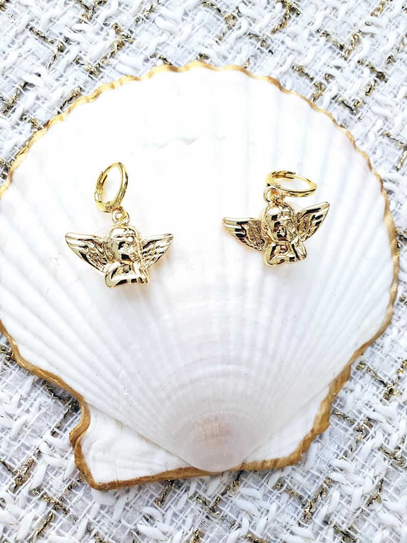 May include: A pair of gold angel-shaped earrings with hoop closures. The earrings are displayed on a white seashell.