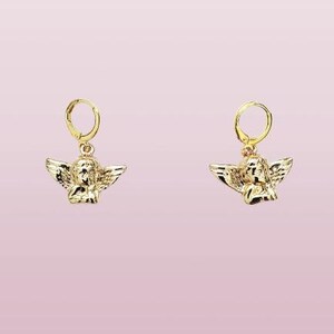 May include: A pair of gold hoop earrings with a small angel charm hanging from each hoop. The angel charms are detailed and have wings.