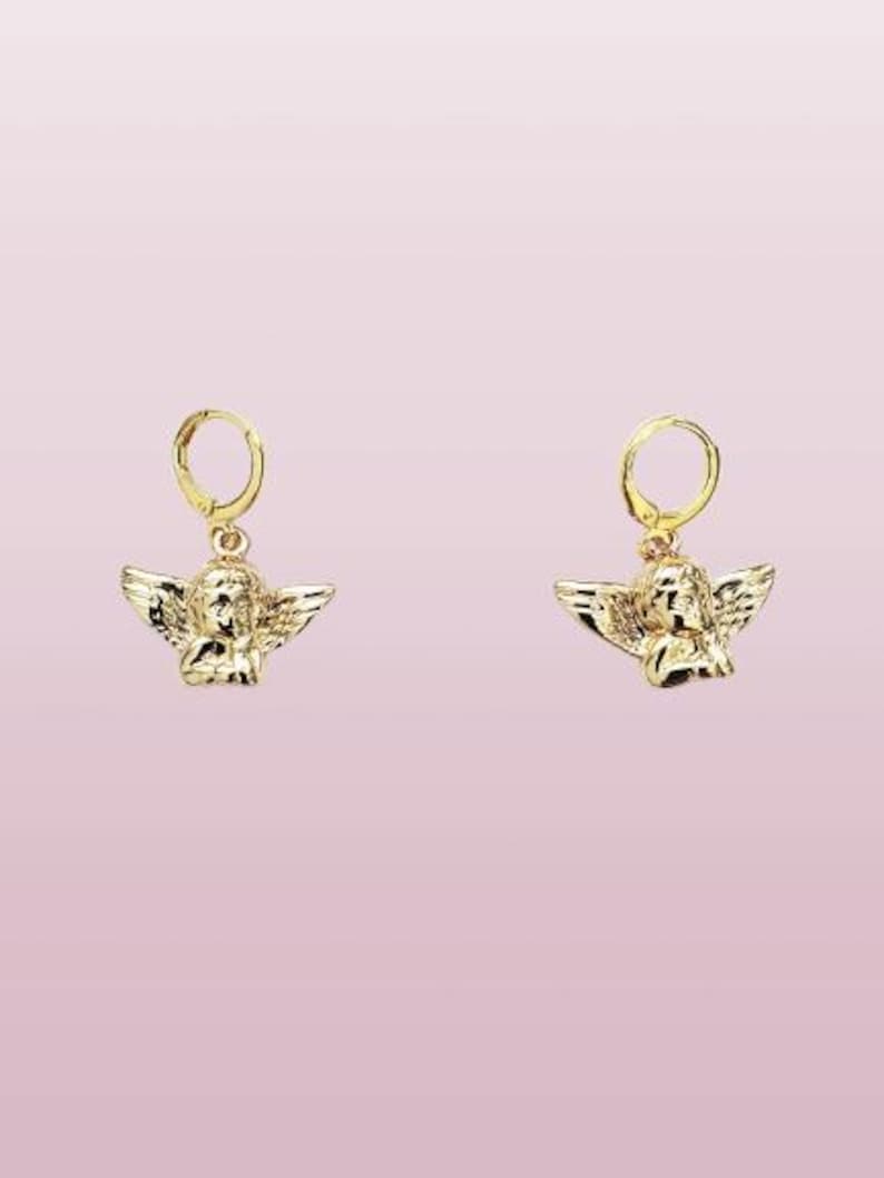 May include: A pair of gold hoop earrings with a small angel charm hanging from each hoop. The angel charms are detailed and have wings.