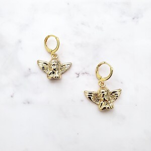 May include: A pair of gold hoop earrings with a small angel charm hanging from each hoop.