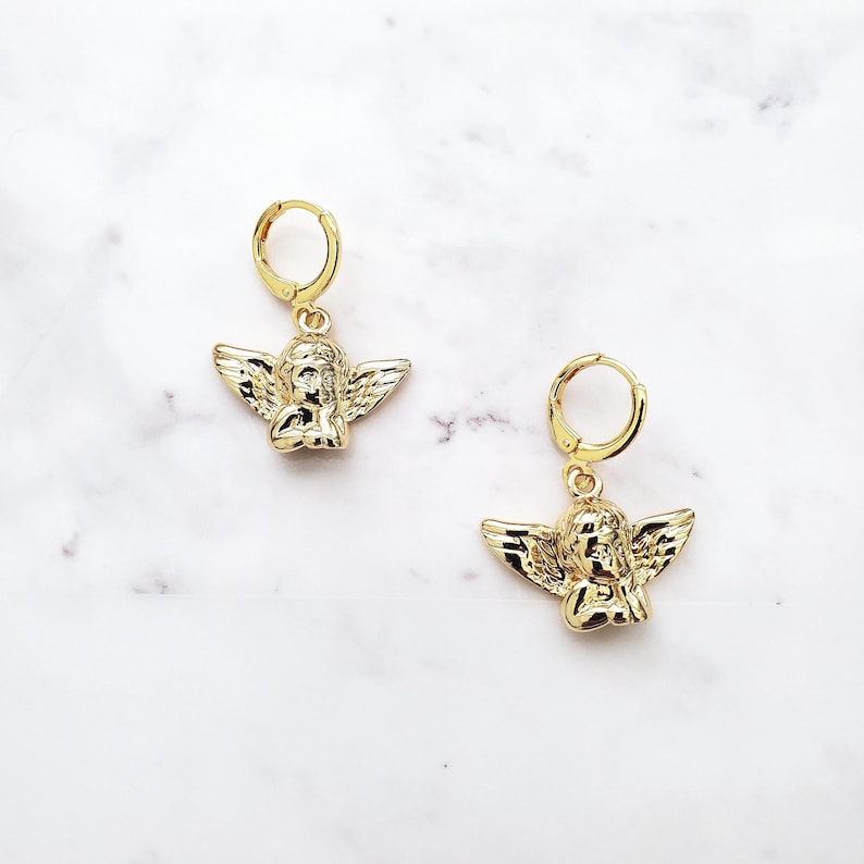 May include: A pair of gold hoop earrings with a small angel charm hanging from each hoop.