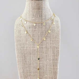 May include: A gold layered necklace with small round charms. The necklace is on a fabric-covered mannequin.