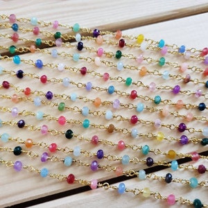May include: A gold-toned metal chain featuring a series of small, faceted, multi-colored gemstone beads spaced evenly along the links. The beads display a variety of hues including pink, blue, green, yellow, purple, and white, creating a vibrant, repeating pattern throughout the length of the delicate jewelry chain.