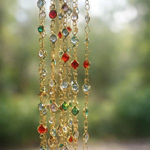May include: A collection of gold-toned chains featuring bezel-set faceted crystals in various colors including red, green, light blue, and clear. The delicate metal links are arranged vertically, showcasing a repeating pattern of colorful stones.