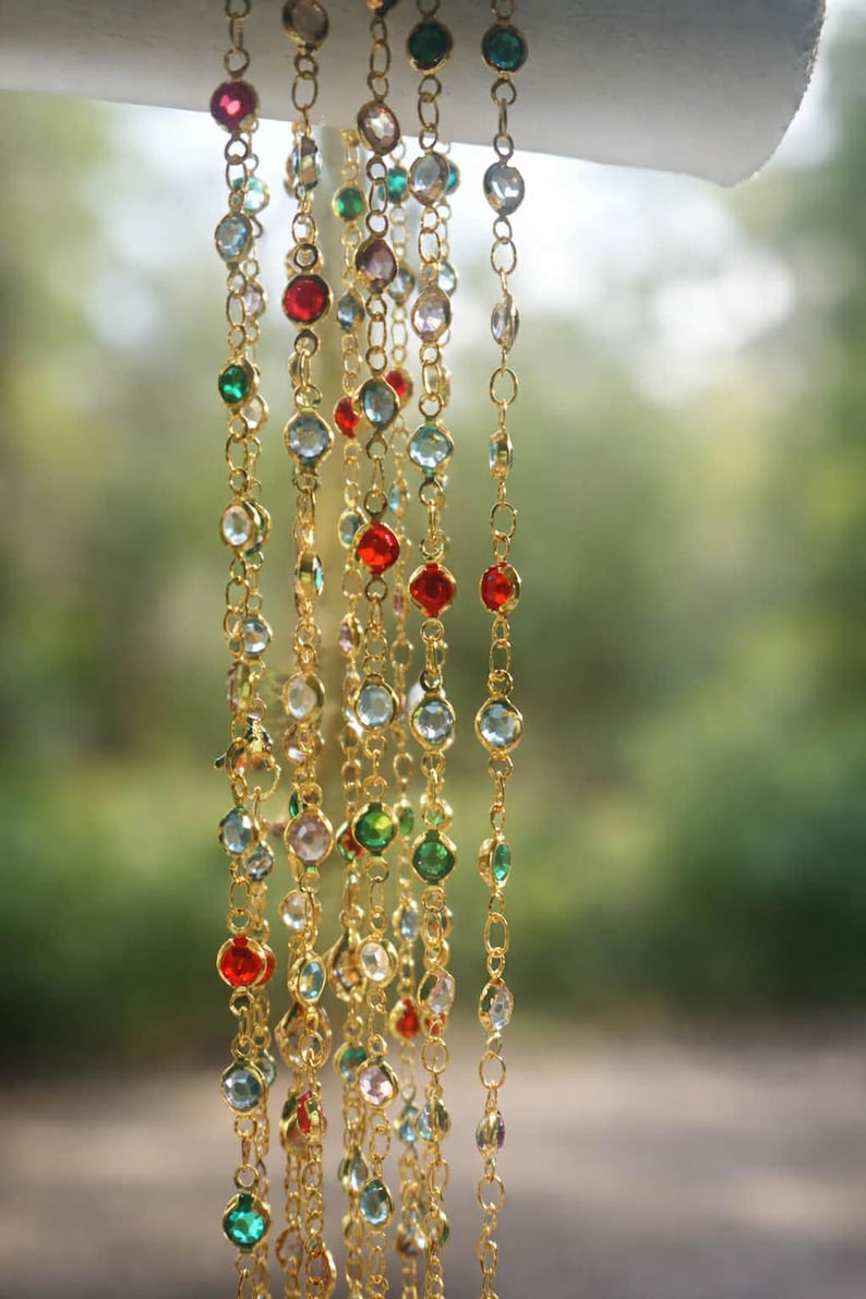 May include: A collection of gold-toned chains featuring bezel-set faceted crystals in various colors including red, green, light blue, and clear. The delicate metal links are arranged vertically, showcasing a repeating pattern of colorful stones.