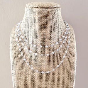 May include: A layered necklace with three strands of small, clear beads. The beads are strung on a delicate silver chain. The necklace is displayed on a burlap-covered bust, against a neutral background.