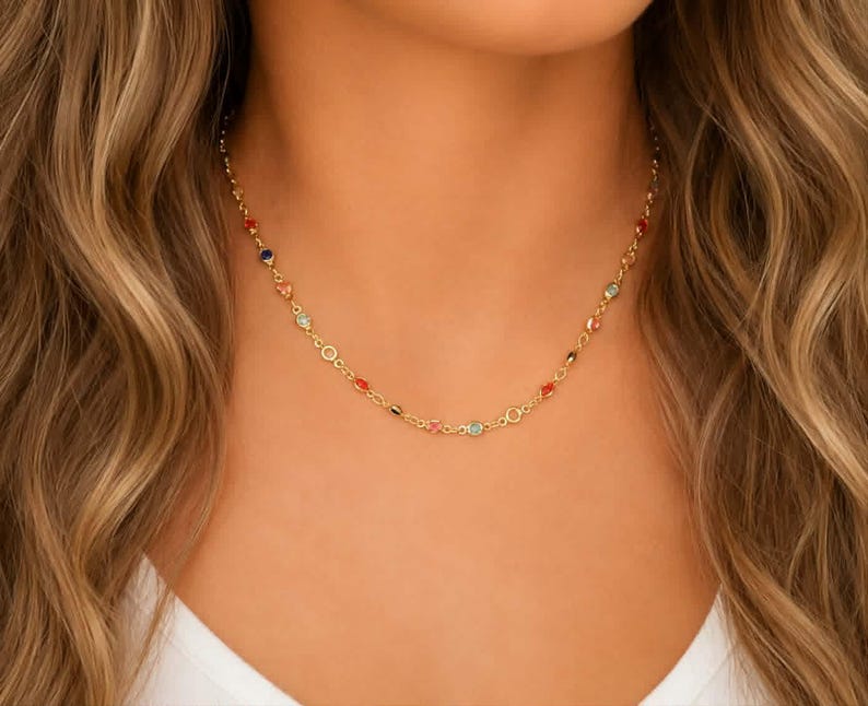 May include: A gold chain necklace featuring small, evenly spaced, multi-colored gemstones in shades of red, blue, and light green. The delicate metal links connect each stone, creating a continuous, lightweight design that rests around the neck.