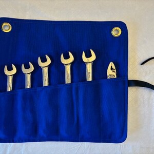May include: A blue canvas tool roll with six wrench pockets and a separate pocket for pliers. The roll has two brass eyelets and a black strap.