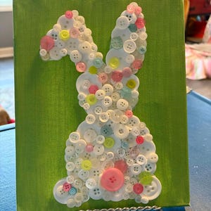 May include: A canvas with a green background features a white bunny silhouette crafted from various sizes of white, pink, yellow, and blue buttons. The bunny is centered on the canvas, creating a whimsical Easter-themed artwork.