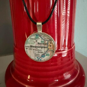 May include: A round pendant necklace with a map design, featuring the text "Normal Bloomington". The pendant is set in a silver-colored bezel and hangs from a black cord. The background is a red, glossy vase.