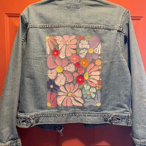 May include: A light-wash denim jacket with a floral design on the back. The design includes various flowers in pink, purple, and orange, with text such as "My Body, My Choice" and "Black Lives Matter".