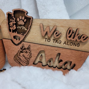 May include: Wooden sign with the text "We like to tag along" and the name "Aska" in a cursive font. A wolf silhouette is carved into the wood. The sign is made of two pieces of wood, one light brown and one dark brown.
