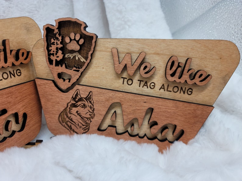 May include: Wooden sign with the text "We like to tag along" and the name "Aska" in a cursive font. A wolf silhouette is carved into the wood. The sign is made of two pieces of wood, one light brown and one dark brown.