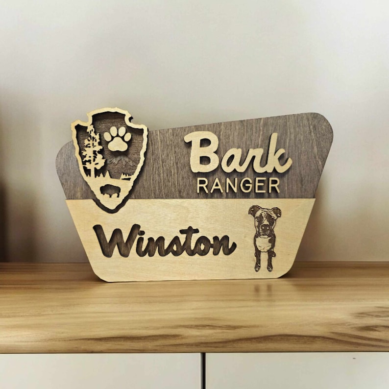 May include: A wooden sign with a brown and light wood finish. The sign has a cutout of a park ranger badge with a paw print and the text "Bark Ranger" above the text "Winston". A black and white photo of a dog is below the text "Winston".