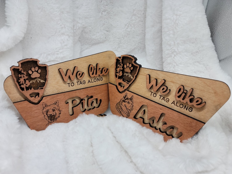 May include: Two wooden signs with the text "We like to tag along" and the names "Pita" and "Aska". Each sign features a wood-burned image of a dog and a national park emblem.