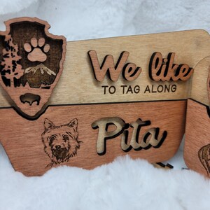 May include: Wooden sign with a paw print and a mountain scene. The sign says "We like to tag along" and "Pita".