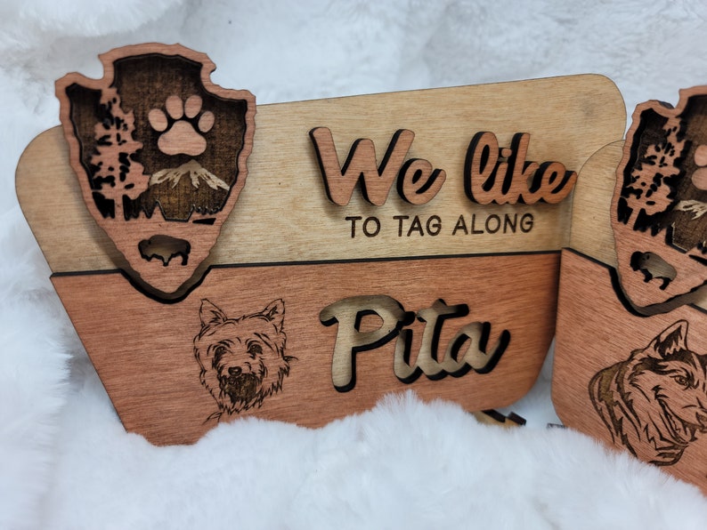 May include: Wooden sign with a paw print and a mountain scene. The sign says "We like to tag along" and "Pita".