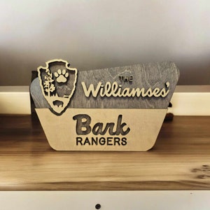 May include: A wooden sign with a brown and tan finish. The sign is shaped like a park ranger's badge and says "The Williamses' Bark Rangers" in a stylized font.