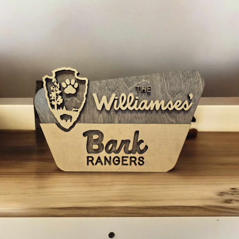 May include: A wooden sign with a brown and tan finish. The sign is shaped like a park ranger's badge and says "The Williamses' Bark Rangers" in a stylized font.
