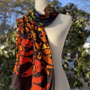 May include: A lightweight scarf featuring a repeating pattern of dancing bears in vibrant shades of orange, yellow, green, and blue set against a dark brown background. The scarf is draped over a white mannequin torso, displaying the colorful graphic print.