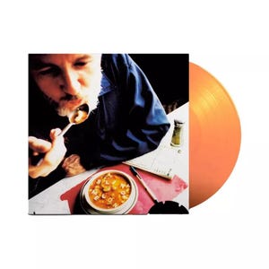 May include: Album cover featuring a man eating soup, with the words "Blind Melon" spelled out in the soup. An orange vinyl record is visible on the right side of the image. A salt shaker and pen are also on the table.