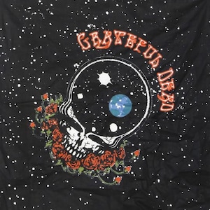 May include: A black Grateful Dead tapestry with a white, starry sky background. The Grateful Dead logo is in orange and the band's iconic skull and roses design is in white. The skull has a blue and green planet inside of it.