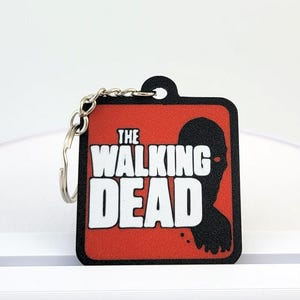 May include: Square keychain featuring the text "THE WALKING DEAD" in white on a red background. A black silhouette of a person is on the right side. The keychain has a silver keyring attached.