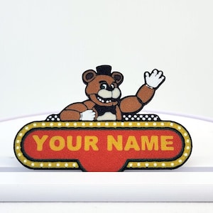 Peut inclure: Un personnage d'ours brun avec un chapeau haut de forme noir et un nœud papillon salue depuis une enseigne rouge qui dit "YOUR NAME". L'enseigne a une bordure jaune avec de petites lumières. L'enseigne est sur une base blanche.