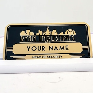 Könnte beinhalten: Ein rechteckiges Namensschild im schwarz-goldenen Art-Déco-Design mit der Aufschrift RYAN INDUSTRIES, YOUR NAME und HEAD OF SECURITY. Das Namensschild wird von einem weißen Kunststoffständer mit einem horizontalen Schlitz aufrecht gehalten.