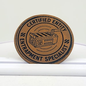 Può includere: Una toppa rotonda in pelle marrone con la scritta "CERTIFIED ENTITY ENTRAPMENT SPECIALIST" che circonda un disegno di trappola. La toppa è esposta su un supporto bianco.