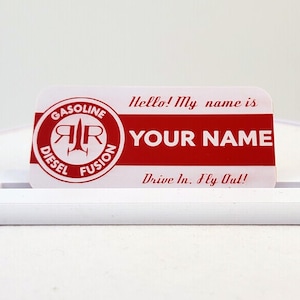 Peut inclure: Un badge nominatif rectangulaire au design rouge et blanc portant le texte Hello! My name is, YOUR NAME, et Drive In. Fly Out! Un logo circulaire à gauche affiche le texte GASOLINE DIESEL FUSION avec un graphique de fusée stylisé.
