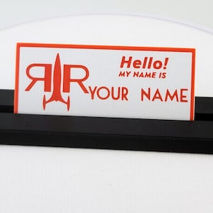 May include: A black name card holder with a white card featuring red text and a rocket ship logo. The card reads "Hello! MY NAME IS YOUR NAME."