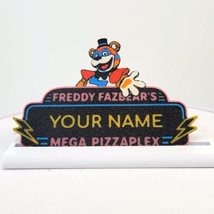 May include: A rectangular desk nameplate featuring a black background with the text FREDDY FAZBEAR'S, YOUR NAME, and MEGA PIZZAPLEX. A cartoon bear character with blue and red accents is positioned on top, flanked by two yellow lightning bolt icons.