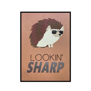 May include: A whimsical illustration of a hedgehog wearing sunglasses, set against a brown background. The text "LOOKIN' SHARP" is displayed in silver lettering below the hedgehog. The image is framed in black.