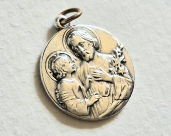 May include: A round silver-toned metal pendant featuring a raised relief design of an adult figure with a beard and a child, both with halos. The adult holds a flowering branch in one hand and rests the other on their chest. A small metal jump ring is attached to the top.
