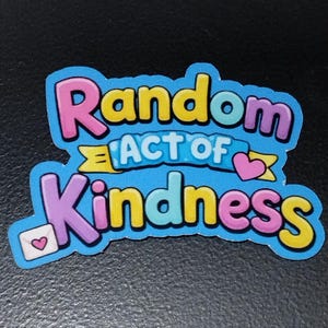 May include: A colorful sticker with the words "Random Act of Kindness" in bubble letters. The sticker has a blue background with pink, yellow, and purple text. A small envelope with a heart is in the lower left corner.