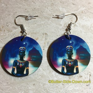 May include: A pair of round wooden earrings with a blue, purple, and pink background featuring a black woman wearing a gold crown and a gold necklace. The woman is sitting in front of a pyramid with stars in the background.