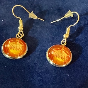 Puede incluir: Un par de pendientes colgantes con marcos de metal plateado. Cada pendiente presenta una imagen redonda, de color naranja con un diseño blanco. Los pendientes tienen un cierre de gancho con un tapón de plástico transparente.