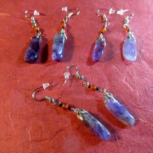 May include: Five pairs of earrings with amethyst stone drops and silver accents. The earrings are on a red background.