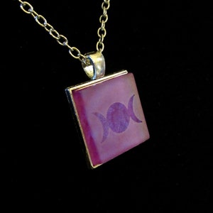 Puede incluir: Collar plateado con un colgante cuadrado. El colgante presenta un símbolo de triple luna púrpura sobre un fondo rosa, engastado en un marco plateado. La cadena es de estilo eslabón delicado, adecuado para el uso diario.