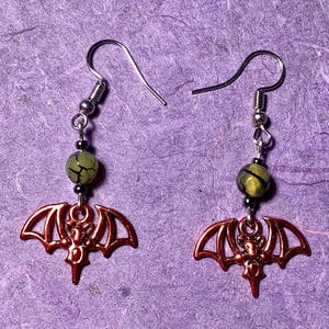May include: A pair of silver bat earrings with green and black agate beads. The bats are a copper color and have a detailed design.