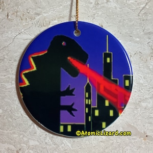 May include: A round ceramic ornament featuring a black silhouette of Godzilla-like monster breathing fire against a purple and blue cityscape. The ornament has a gold string for hanging and the text "AtomicLizard.com" at the bottom.