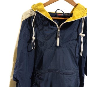 Puede incluir: Chaqueta anorak azul marino y amarilla con cremallera y cordón blancos. La chaqueta tiene un bolsillo delantero y una capucha amarilla. La marca "WINNER" es visible en la cremallera y la etiqueta.